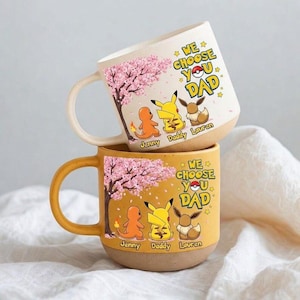 May include: Two ceramic mugs, one on top of the other. The top mug is off-white, the bottom is mustard yellow. Both have a cherry blossom design, Pokemon characters, and the text "WE CHOOSE YOU DAD".