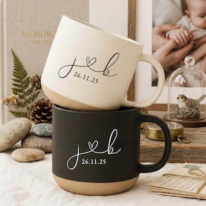 May include: Two stacked ceramic mugs, one cream and one black, with a tan base. Each mug features the initials "jeb", a heart, and the date "26.11.25" in a delicate script. The mugs have a rounded shape and a large handle.