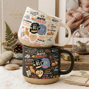 Puede incluir: Dos tazas de cerámica con bases blancas y negras. La taza superior es blanca con una base color crema, y la taza inferior es negra con una base color crema. Ambas tazas presentan el texto "HEATED RIVALRY" y gráficos de camisetas de hockey. Las tazas están decoradas con varias ilustraciones y frases.