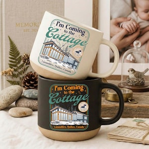 May include: Two ceramic mugs, one cream-colored and one black, stacked on top of each other. Both mugs feature the text "I'm Coming to the Cottage" with a graphic of a cottage and the location "Lanaudière, Québec, Canada."
