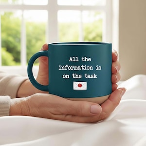 May include: A teal ceramic mug with a tan base, featuring the text "All the information is on the task" and an envelope graphic. The mug has a curved handle and is held by a person.