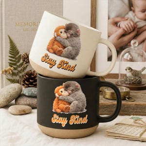 May include: Two ceramic mugs stacked on top of each other. The top mug is off-white with a brown base, and the bottom mug is black with a brown base. Each mug features a graphic of two hugging monkeys and the text "Stay Kind".