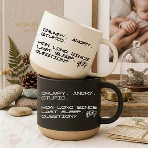 May include: Two ceramic mugs, one cream and one black, stacked on top of each other. The cream mug has a tan base and black text that reads "CRUMPY. STUPID. ANGRY. HOW LONG SINCE LAST SLEEP? QUESTION?". The black mug has the same text.