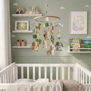 May include: A nursery mobile featuring felted plush rabbits in shades of brown and gray, suspended from a circular wooden frame with felt clouds, carrots, flowers, and green leaves hanging on strings above a white wooden crib.