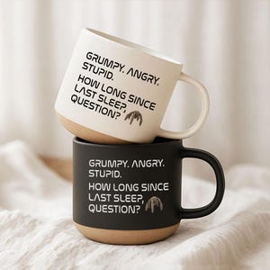 May include: Two ceramic mugs, one white and one black, stacked on top of each other. Each mug has a brown base and a curved handle. The white mug has the text "GRUMPY. ANGRY. STUPID. HOW LONG SINCE LAST SLEEP, QUESTION?" printed in black. The black mug has the same text in white.
