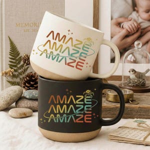 Puede incluir: Dos tazas de cerámica apiladas. La taza superior es blanquecina, la inferior es negra. Ambas tazas tienen la palabra "AMAZE" en una fuente retro con un diseño de planeta. Las tazas tienen una base beige.