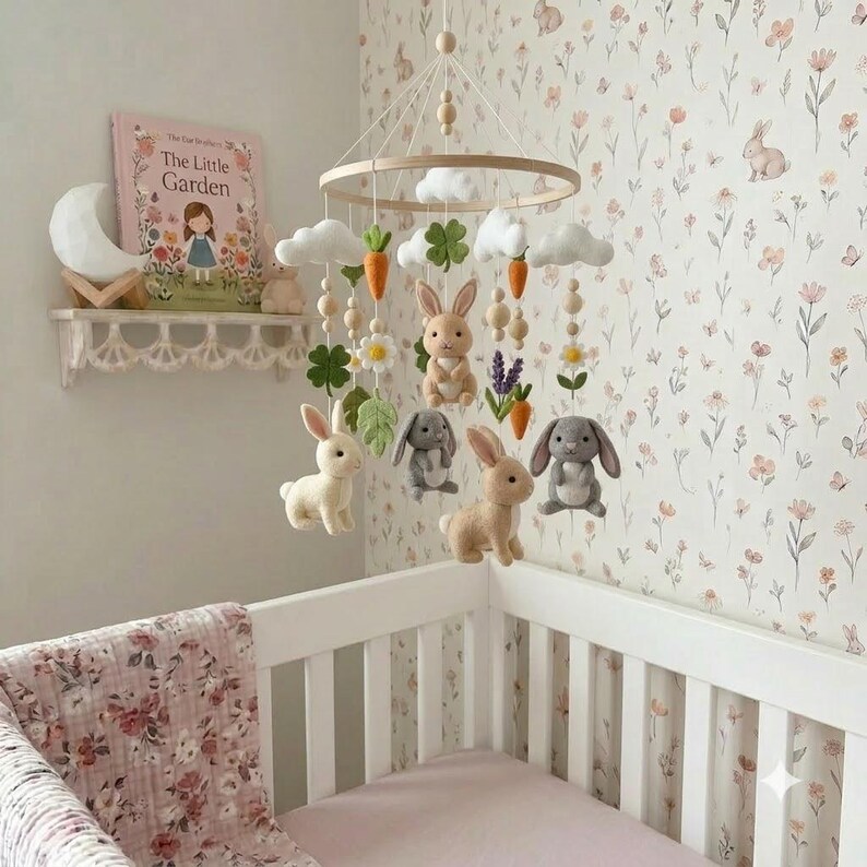 May include: A nursery mobile featuring felted figures of rabbits in white, beige, and gray, suspended from a circular wooden frame. The mobile includes felt clouds, orange carrots, green clover leaves, and white flowers, all hanging from strings with small wooden beads.