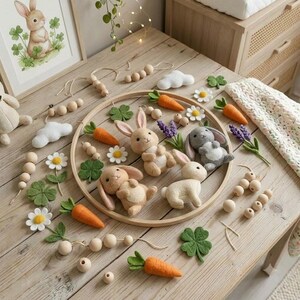 May include: A circular wooden hoop mobile featuring three felt rabbits in tan, cream, and gray, surrounded by felt carrots, white daisies, green clover leaves, purple lavender sprigs, white clouds, and natural wooden beads on strings.