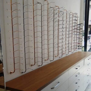 Pode incluir: Um expositor de parede com v&aacute;rias hastes met&aacute;licas verticais em acabamentos de cobre e prata, equipadas com ganchos horizontais para organizar &oacute;culos. Abaixo do expositor, h&aacute; um arm&aacute;rio branco com um tampo de madeira e puxadores.