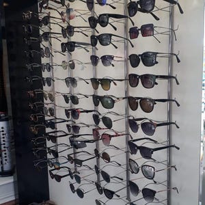 Pode incluir: Um expositor de parede com numerosos pares de &oacute;culos de sol e &oacute;culos de grau em v&aacute;rios estilos, cores de arma&ccedil;&atilde;o e tons de lentes. Os &oacute;culos est&atilde;o organizados em colunas verticais num painel de cor clara com suportes de metal.