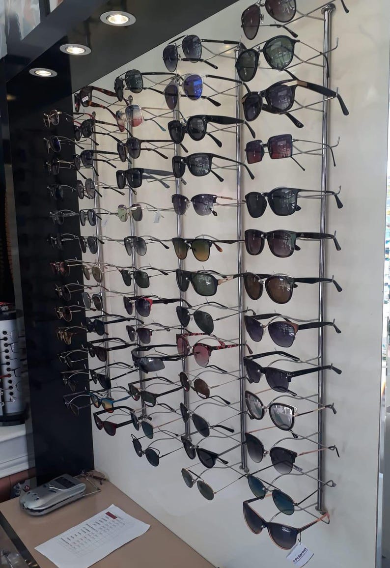 Pode incluir: Um expositor de parede com numerosos pares de &oacute;culos de sol e &oacute;culos de grau em v&aacute;rios estilos, cores de arma&ccedil;&atilde;o e tons de lentes. Os &oacute;culos est&atilde;o organizados em colunas verticais num painel de cor clara com suportes de metal.