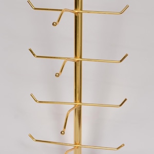 Pode incluir: Um expositor vertical de metal dourado com uma haste central e v&aacute;rios bra&ccedil;os horizontais que se estendem para fora. Cada bra&ccedil;o termina com uma pequena ponta arredondada, projetada para segurar itens com seguran&ccedil;a nesta estrutura met&aacute;lica.