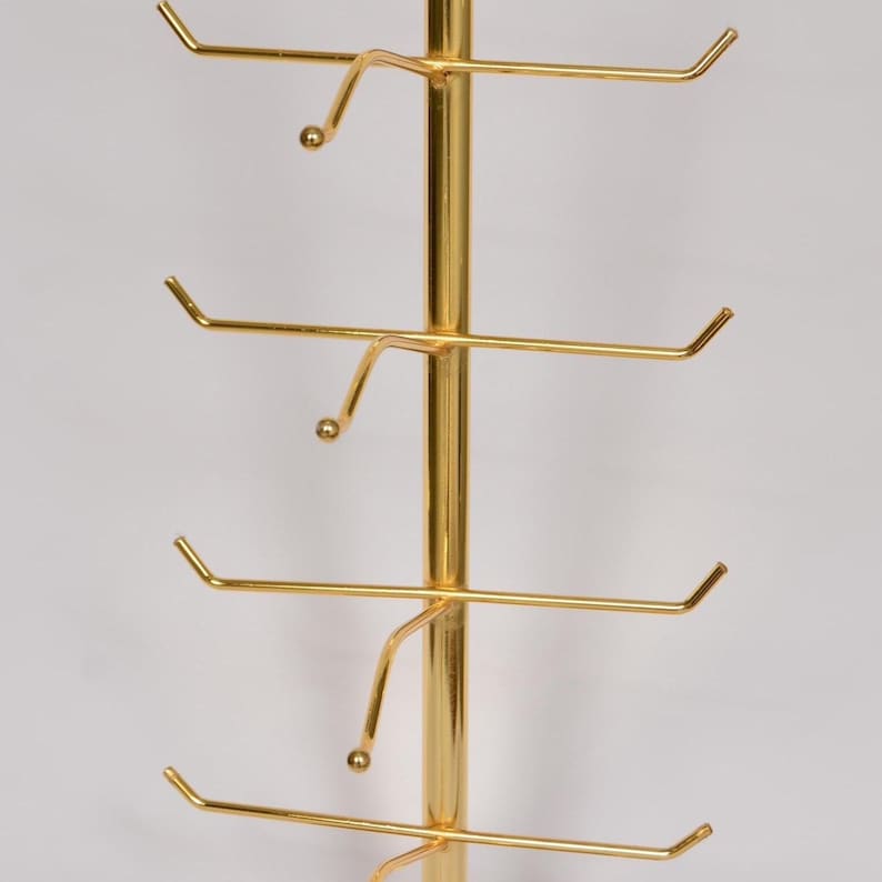 Pode incluir: Um expositor vertical de metal dourado com uma haste central e v&aacute;rios bra&ccedil;os horizontais que se estendem para fora. Cada bra&ccedil;o termina com uma pequena ponta arredondada, projetada para segurar itens com seguran&ccedil;a nesta estrutura met&aacute;lica.