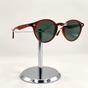 Può includere: Occhiali da sole rotondi con montatura color ambra traslucido e lenti verde scuro. Gli occhiali presentano cerniere e aste in metallo color argento, poggiati su un espositore in cromo lucido con base circolare.
