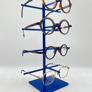 Peut inclure: Un présentoir vertical en métal bleu avec quatre bras horizontaux supportant quatre paires de lunettes. La paire supérieure a un motif écaille, la deuxième est violet foncé, la troisième est écaille marron et la paire inférieure est dorée.