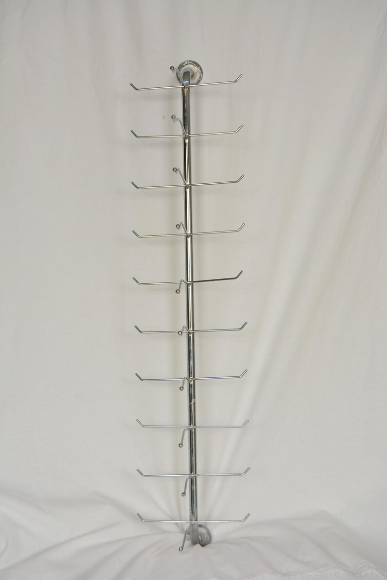 Pode incluir: Um expositor vertical de metal prateado com uma haste central e dez bra&ccedil;os horizontais que se estendem de cada lado. A estrutura inclui um suporte de montagem circular no topo e uma base curva na parte inferior para estabilidade.