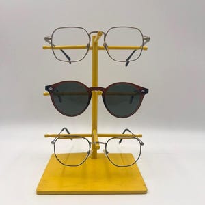 Peut inclure: Un présentoir à lunettes en métal jaune avec trois paires de lunettes. La paire supérieure est cerclée d'argent et de forme géométrique. La paire du milieu est des lunettes de soleil marron. La paire inférieure est des lunettes à monture noire.