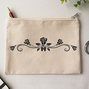 May include: A beige canvas zipper pouch with a black floral design. The design features roses and leaves in a symmetrical pattern. A red pen is visible in the open pouch. The pouch is rectangular and has a zipper closure.