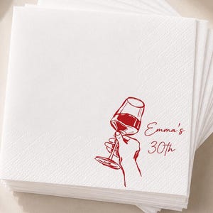 May include: A stack of square white paper napkins featuring a red line art illustration of a hand holding a wine glass. The text "Emma's 30th" is printed in red script next to the illustration. The napkins have a textured border pattern.