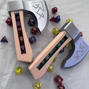 May include: Two novelty dice holders shaped like axes, one peach and one lavender, each with a silver blade and a compartment filled with dice. Assorted polyhedral dice in various colors are scattered around the axes.