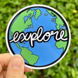 May include: A round sticker featuring a blue and green Earth graphic with the word "explore" in white script. The sticker has a black border and is held against a blurred green background.