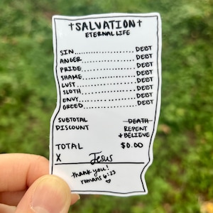 May include: A white sticker with black text, resembling a receipt, held against a blurred green background. The sticker lists sins and debt, with "Salvation" at the top. The total is $0.00, with "Jesus" and a Bible verse at the bottom.