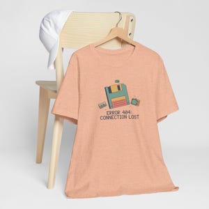 Op de afbeelding: Perzikkleurig T-shirt met een retro computergrafiek met een diskette, cassettebandje en een klein scherm. De tekst "ERROR 404: CONNECTION LOST" staat eronder gedrukt. Een witte pet ligt op een houten stoel.