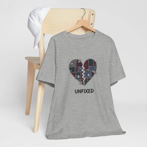 Peut inclure: T-shirt gris avec un motif de cœur brisé en patchwork. Le cœur est divisé en deux par un motif en zigzag. Le mot "UNFIXED" est imprimé en noir en dessous.