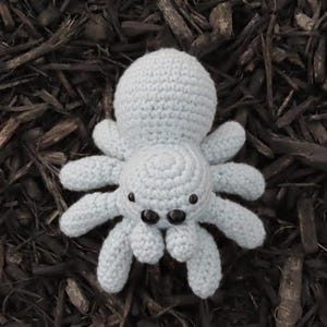 Puede incluir: Una araña de ganchillo azul claro con ocho patas y dos ojos negros. La araña hecha a mano descansa sobre un lecho de mantillo marrón oscuro. El cuerpo y las patas de la araña están hechos de hilo suave.