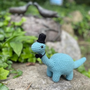 Puede incluir: Un dinosaurio de ganchillo azul claro con sombrero de copa negro y pajarita. El juguete amigurumi hecho a mano está sobre una roca, con un fondo verde borroso.
