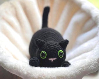 May include: A handmade black crochet cat with large green eyes and a pink nose, resting on a fluffy white surface. The cat has small ears and a curled tail. The cat is a cute, handmade item.