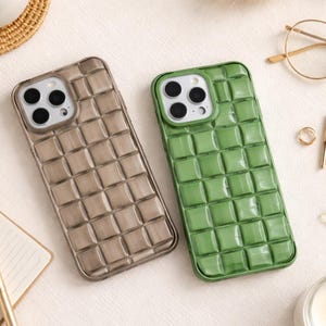 Puede incluir: Dos fundas de teléfono con diseño acolchado. Una es marrón claro, la otra verde. Ambas tienen un patrón cuadrado y están diseñadas para un smartphone. Las fundas se muestran sobre una superficie blanca.