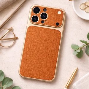 Può includere: Custodia per telefono arancione con cornice dorata e finitura strutturata. La custodia ha una sezione per gli obiettivi della fotocamera. Lo sfondo include una penna, occhiali e una piccola pianta.