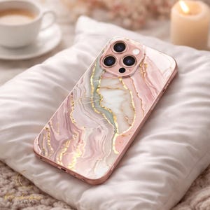Puede incluir: Funda de teléfono con diseño de mármol rosa y dorado. La funda tiene un borde de oro rosa y un diseño que se asemeja a mármol arremolinado con detalles dorados. La funda está sobre una almohada blanca.