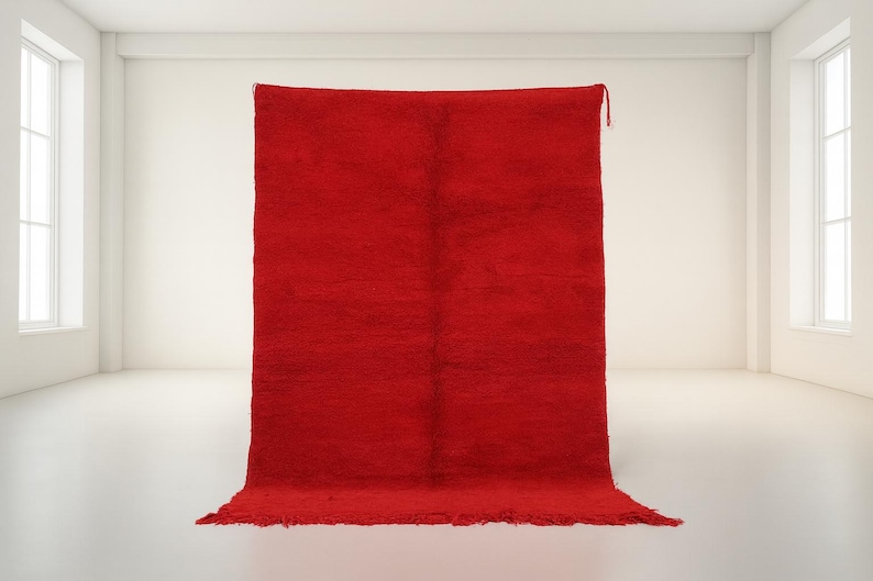A large, vibrant red rug is displayed in a minimalist, white-walled room with two windows.