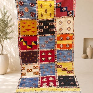 Peut inclure: Un tapis rectangulaire composé d'une grille de carrés et de rectangles multicolores. Chaque section présente des motifs géométriques distincts, notamment des losanges, des triangles et des fleurs dans des tons bleu, jaune, rouge, noir et crème.