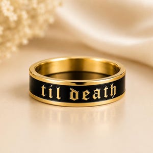 May include: A gold-toned band ring featuring a black enamel inlay that wraps around the center. The words "til death" are engraved in a gothic-style font across the black surface, with the gold metal visible on the edges and the interior.