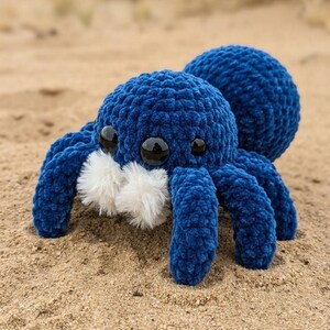 May include: A plush, blue crocheted spider toy with eight legs, two large eyes, and white fluffy accents. The spider is resting on a sandy surface, with a blurred background of sand and vegetation.