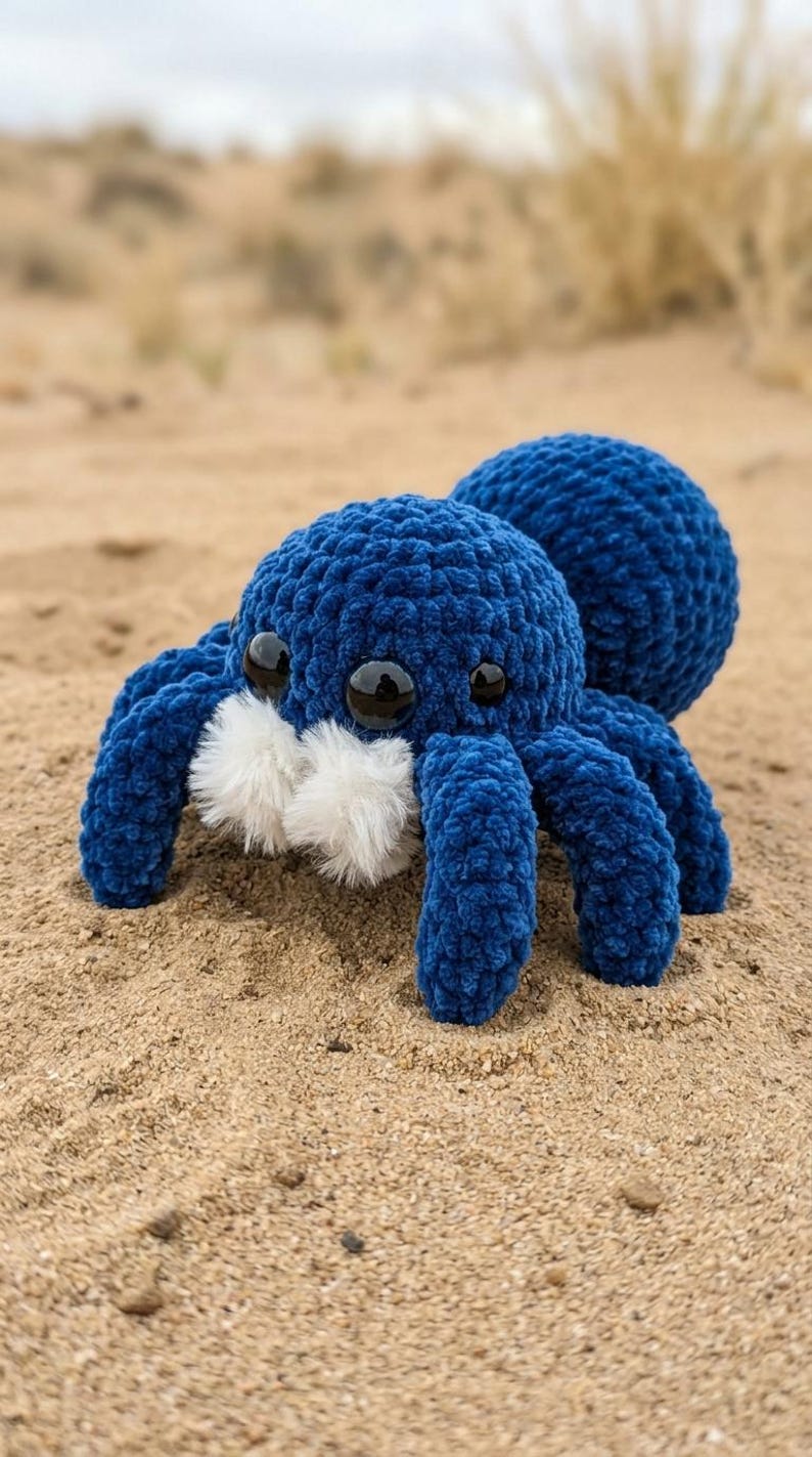May include: A plush, blue crocheted spider toy with eight legs, two large eyes, and white fluffy accents. The spider is resting on a sandy surface, with a blurred background of sand and vegetation.
