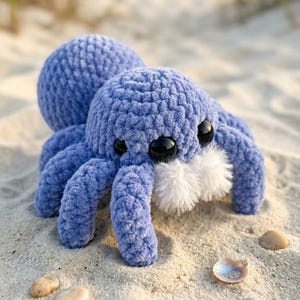 May include: A plush, blue spider toy with large black eyes and a fluffy white beard. The crocheted spider is resting on a sandy beach with small seashells scattered around it. The spider's body is a soft, textured material.
