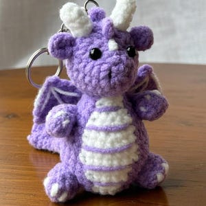 Könnte beinhalten: Ein handgefertigter Drachen-Schlüsselanhänger aus Häkelarbeit in Lila und Weiß. Der Drache hat ein detailliertes Design mit Flügeln, Hörnern und einem gestreiften Bauch. Der Schlüsselanhänger liegt auf einer Holzoberfläche.