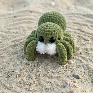 May include: A handmade, green crocheted spider with large black eyes and fluffy white accents. The plush toy is resting on a sandy surface with small pebbles and seashells.