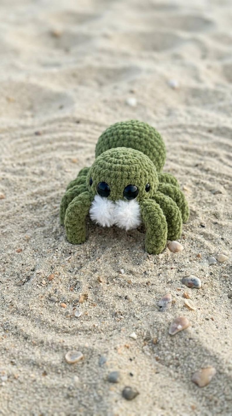 May include: A handmade, green crocheted spider with large black eyes and fluffy white accents. The plush toy is resting on a sandy surface with small pebbles and seashells.