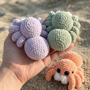 May include: Three crocheted spiders in shades of lavender, green, and peach with black eyes and white accents. The handmade toys are displayed on a sandy beach, showcasing their soft, textured bodies and eight legs.