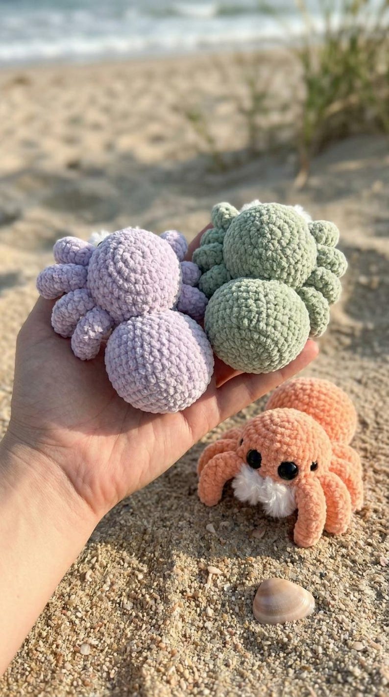 May include: Three crocheted spiders in shades of lavender, green, and peach with black eyes and white accents. The handmade toys are displayed on a sandy beach, showcasing their soft, textured bodies and eight legs.