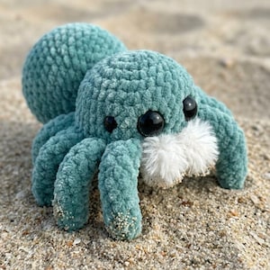 Puede incluir: Un peluche de araña de ganchillo turquesa con grandes ojos negros y una boca blanca y esponjosa. El peluche descansa sobre una playa de arena, con el océano y las dunas al fondo.