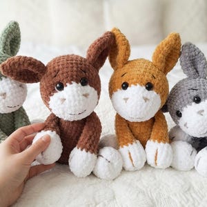 Puede incluir: Cuatro peluches de burro hechos a mano con ganchillo en tonos verdes, marrones, naranjas y grises. Cada burro tiene detalles blancos en la cara y los pies, con orejas grandes y una expresión sonriente. Los juguetes están colocados sobre una colcha blanca.
