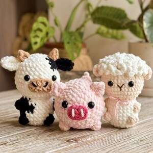 Könnte beinhalten: Drei gehäkelte Bauernhoftierfiguren: eine schwarz-weiße Kuh, ein rosa Schwein und ein weißes Schaf. Die Kuh hat schwarze Flecken und beige Ohren. Das Schwein hat eine rosa Schnauze. Das Schaf hat einen flauschigen weißen Kopf und eine rosa Schleife.