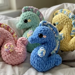 Puede incluir: Cuatro peluches de caballitos de mar hechos a mano con ganchillo, en colores pastel: rosa, verde menta, azul y amarillo. Cada uno tiene una melena detallada y grandes ojos negros, sobre una superficie blanca y suave.