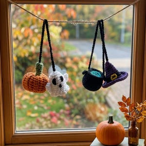 Könnte beinhalten: Ein Set aus vier gehäkelten Halloween-Anhängern, die an einer Schnur hängen, bestehend aus einem orangefarbenen Kürbis, einem weißen Geist, einem schwarzen Kessel mit grüner Flüssigkeit und einem violetten Hexenhut mit gelber Schnalle sowie einem Kürbis.